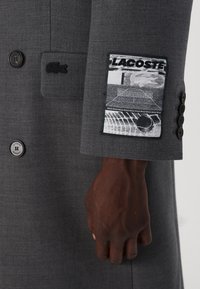 Cappotto di lana grigio con chiusura a bottoni, tessuto testurizzato e un grande patch del logo Lacoste in bianco e nero sulla manica.