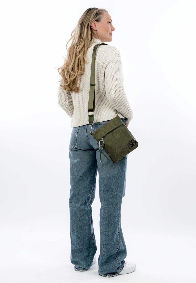 Olive green crossbody bag made of textured fabric, featuring a zip closure and adjustable strap, worn over a cream sweater and blue jeans.