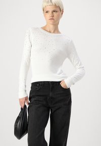 Pulover alb cu mâneci lungi, cu guler cusut în ondulă, decorat cu cuie argintii, asortat cu blugi negri din denim cu talie înaltă și o geantă neagră.