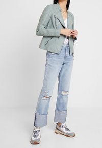 Lichtgroene suède motorjack met geribbelde details, gecombineerd met versleten lichtblauwe jeans met een rafelige zoom en grijze sneakers.
