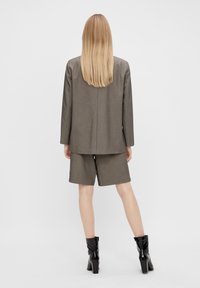 Blazer marron avec short assorti, tous deux en tissu texturé. Le modèle porte des bottines noires à talons, cheveux lâchés, dos tourné à la caméra.