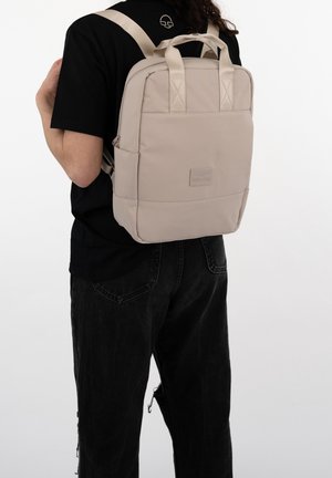 Sac à dos rectangulaire beige avec bretelles rembourrées et poignée supérieure. Présente une texture lisse, une poche zippée à l'avant et un patch logo à la base.