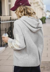 Gray hooded sweater made of soft fabric featuring ribbed cuffs, striped accents, and a relaxed fit, held by a person with a drink.