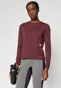 Burgundi pika pikad käised sportlik särk ümmarguse kaelusega, pehme kangas ja logoDETAIL. Kombineeritud hallide retuusidega ja musta veepudeli kanssa.