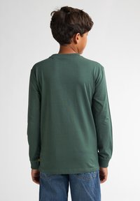 Camicia a maniche lunghe in tessuto verde scuro con colletto rotondo, dotata di orlo dritto e polsini a coste, indossata con jeans blu.