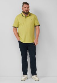 Gelbes Poloshirt mit dunkel gestreiftem Kragen, kombiniert mit dunkelblauen Jeans und hellen Sneakers. Einfaches, klassisches Design mit minimalem Branding.