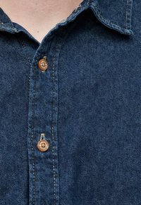 Σκούρο μπλε πουκάμισο από τζιν με κλείσιμο με κουμπιά, με δύο καφέ κουμπιά και ελαφρώς φθαρμένο γιακά. Λείο ύφασμα, casual σχεδιασμός.