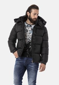 Schwarze Daunenjacke mit Kapuze, Reißverschluss vorne, geprägtem Logo und gerippten Bündchen, getragen über einem bedruckten T-Shirt und blauen Jeans.