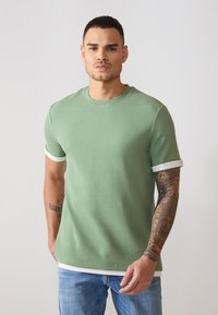 T-shirt vert à manches courtes en maille ribbed avec des poignets et un ourlet rayés blancs. Présente un tissu texturé et un col rond.