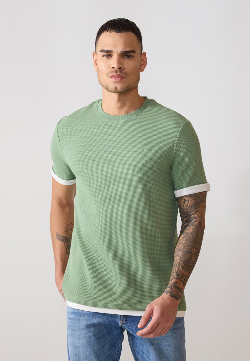 T-shirt vert à manches courtes en maille ribbed avec des poignets et un ourlet rayés blancs. Présente un tissu texturé et un col rond.