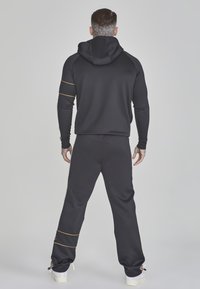 Chándal deportivo negro con capucha, con franjas de acento doradas. Textura suave, diseño ajustado en la parte superior y pantalones sueltos. Zapatillas blancas.