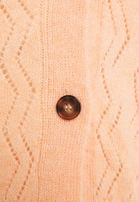 Cardigan en tricot couleur pêche avec un motif en zigzag et un bouton sombre à quatre trous au centre. Texture douce et aspect confortable.