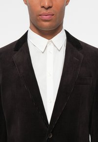 Svart kordfläkt blazer med vertikala ribbor, notch-kragar och en enda bröstficka, bärs över en vit skjorta med knappar.