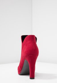 Rode suède enkellaars met hoge hak en taps toelopende vorm, voorzien van een zwarte zool. Soepele textuur, afgeronde randen en minimale stikseldetails.