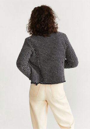 Schwarze und weiße strukturierte, taillierte Jacke mit einem gewebten Muster, fransigen Säumen und langen Ärmeln, getragen über hellen Hosen.