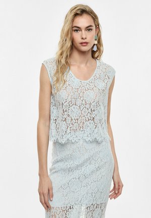 Donna con lunghi capelli biondi indossa un top di pizzo azzurro senza maniche e una gonna coordinata, in piedi davanti a uno sfondo bianco semplice.