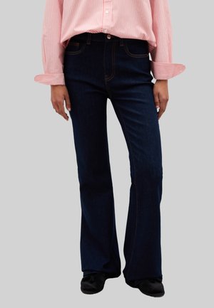 Jeans a zampa d'elefante di un blu scuro con cuciture gialle, vita alta e design a cinque tasche. Indossati con una camicia a maniche lunghe a righe rosa e bianche.