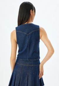 Denimowy crop top o dopasowanym kroju, w ciemnoniebieskim kolorze, bez rękawów, z widocznymi detalami szwów wzdłuż dekoltu i dołu.