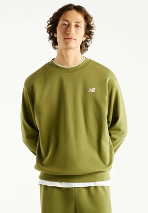Junge Person mit lockigem Haar trägt ein olivgrünes New Balance Sweatshirt und passende Hose, steht mit den Händen hinter dem Rücken.
