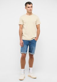 Camiseta básica de algodón color beige claro con cuello redondo, combinada con pantalones cortos de mezclilla azul. Los pantalones cortos están doblados en el dobladillo. Llevado con zapatillas blancas y calcetines con logo.