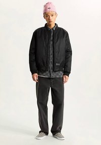 Zwarte bomberjack met geribbelde manchetten, gedragen over een geruit overhemd, gecombineerd met zwarte spijkerbroek en slip-on schoenen met dambordpatroon. Roze gebreide muts als accent.