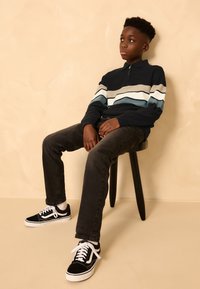 Suéter azul marino con cremallera, con rayas azul claro y crema, combinado con jeans oscuros y zapatillas negras Vans, sentado en un taburete de madera negro.