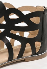 Svart utskuren sandal med en design med intrikata mönster, en slät läderfinish och en beige inredning med platt sula.