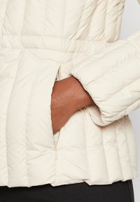 Veste matelassée beige crème avec une poche zippée et des poignets élastiqués. Texture lisse avec des détails de couture horizontale.