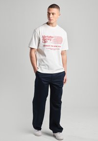 T-shirt blanc avec le texte rouge "Manhattan Running Club", accompagné d'un pantalon bleu marine à coupe ample, assorti à des baskets blanches sur un fond neutre.