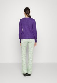 Lila stickad tröja med långa ärmar, kombinerad med utsvängda gröna byxor med ett marmormönster. Svarta skor fullbordar outfiten.