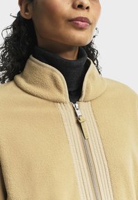 Veste en polaire beige avec un col montant et une fermeture éclair texturée. Portée par-dessus un col roulé gris foncé, avec un détail de fermeture éclair lisse.