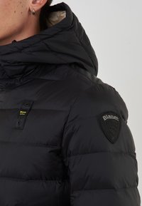 Primer plano de una persona que lleva una chaqueta acolchada negra con capucha, presentada con un parche del logo de Blauer en el hombro y una etiqueta "Blauer U.S.A." en el pecho.
