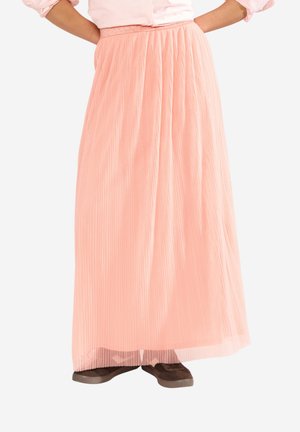 Woman wearing a light pink pleated maxi skirt with a matching blouse and dark brown slip-on shoes, hands resting on hips.