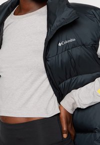Černá péřová vesta s lesklou texturou a logem Columbia, vrstvená přes světle šedou dlouhou halenku a černé kalhoty.