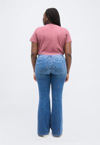 Γυναίκα με πλεξούδες στέκεται με την πλάτη, φορώντας ένα ροζ μπλουζάκι, τζιν καμπάνα μπλε και άσπρα παπούτσια, πάνω σε απλό φόντο.