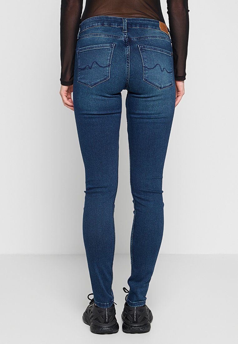 Jeans skinny blu scuro con una texture liscia, dotati di due tasche posteriori con cuciture ondulate e un'etichetta in pelle marrone sulla cintura.