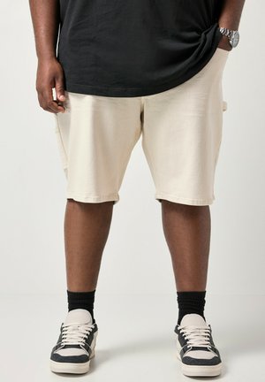 Persoon die beige korte broek draagt, zwarte sokken, zwart-witte sneakers en een zwart t-shirt met één hand in de zak en een horloge aan de andere pols.