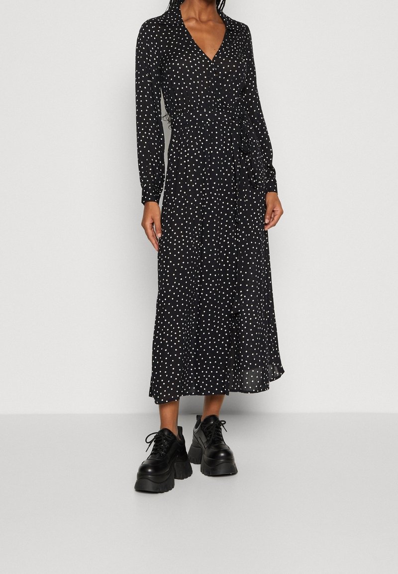 Robe portefeuille noire à pois avec manches longues, encolure en V et ourlet mi-long. Associée à des baskets à plateforme noires épaisses.