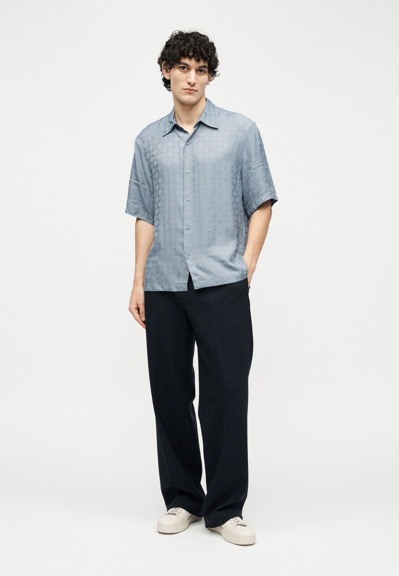 Young man with curly hair wears a light blue short-sleeve button-up shirt, black wide-leg trousers, and white sneakers standing against a white background.