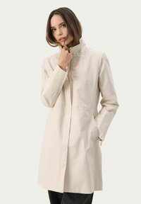 Ljusbeige trenchcoat med hög krage, dragkedja fram, sidofickor och långa ärmar. Slät tyg med en något strukturerad passform.