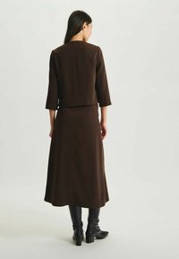 Jupe marron longueur genou avec une veste assortie cintrée. Tissu lisse, manches 3/4, silhouette droite. Portée avec des bottes noires montant au genou.