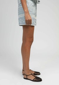 Jambes de femme portant un short léger à motifs bleus clairs et des sandales plates tressées noires avec des brides autour des chevilles sur un fond uni.