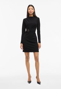 Femeie îmbrăcată cu o rochie neagră lungă, ajustată, cu o cataramă pe partea laterală și cu pantofi cu vârf ascuțit negri, stând pe un fundal alb.