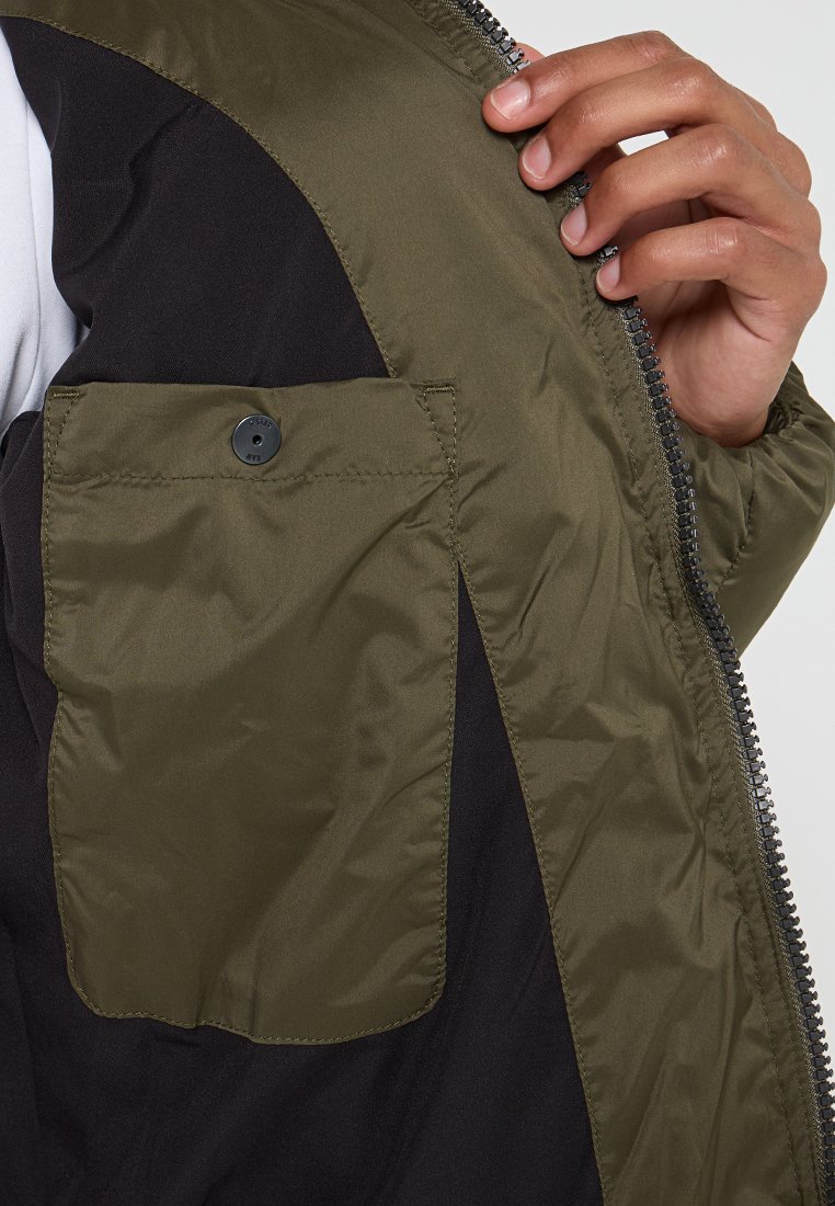 Olivengrüne Jacke mit Reißverschluss, kontrastierendem schwarzen Innenfutter und einer knöpfbaren Brusttasche. Glatte, strapazierfähige Stofftextur.