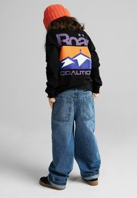 Czarna koszula z długim rękawem z kolorową grafiką gór i napisem "Roar Coalition" na plecach, zestawiona z dużymi niebieskimi dżinsami.