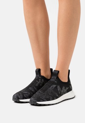 Sneakers neri senza lacci con suole bianche e motivo glitterato a logo indossati su gambe nude su uno sfondo semplice.