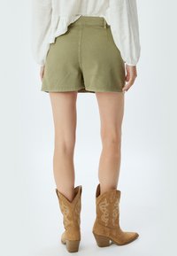 Pantaloni corti verde oliva a taglio dritto, abbinati a stivali da cowboy color crema con cuciture decorative e tacco sottile.