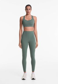 Zeleny športni crop top in legice z visokim pasom, izdelane iz raztegljive tkanine. Model nosi bele superge, kar poudarja prileganje in obliko oblačila.