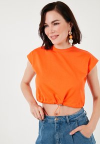 Orange cropped t-shirt med korte ærmer, snøring i kanten og glat bomuldsstruktur, parret med højtaljede blå jeans.