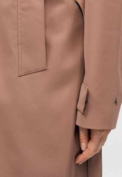 Detailný záber na ruku osoby vedľa manžety hnedej kabátovej bundy s detailom zapínania na gombík.